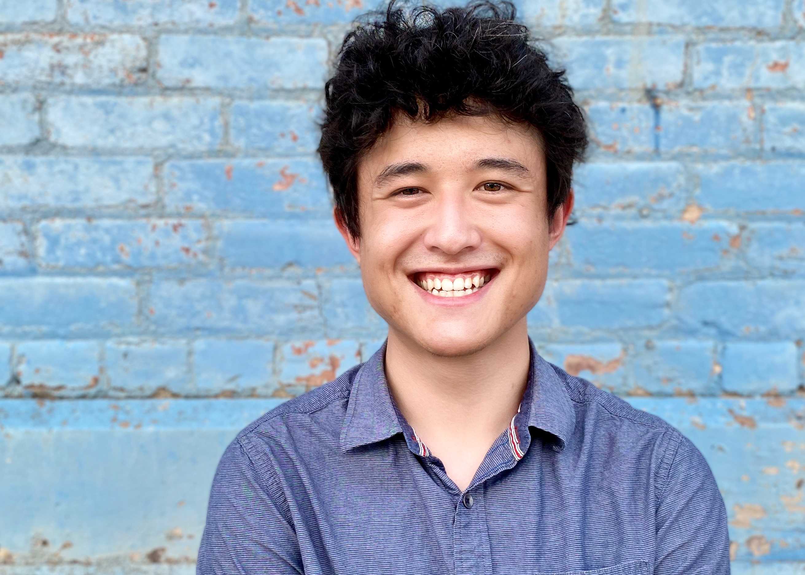 A portrait of Ben Pettis. He is framed from the shoulders up, and is wearing a blue shirt and standing in front of a blue background.